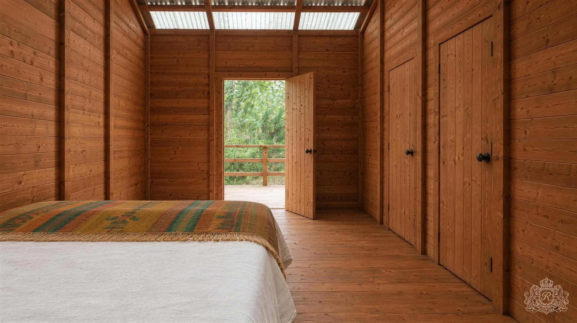 La chambre vue du fond vers l’entrée : lit au drap de lin blanc et plaid tissé à gauche, portes de bois du WC et de la douche à droite, porte ouverte sur la terrasse et sa balustrade