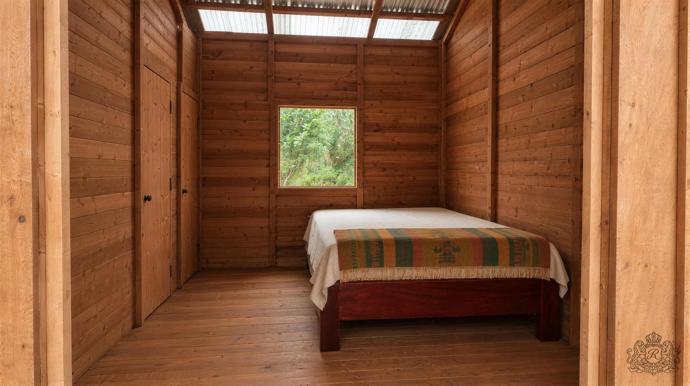 Une chambre du Domaine vue depuis l’entrée : lit de bois, fenêtre carrée, portes de la douche et du WC