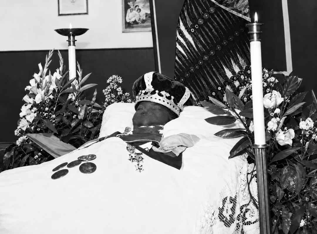 Photographie ancienne en noir et blanc : un chef couronné repose sur un lit drapé de blanc, une décoration sur la poitrine et des médailles posées sur le drap, entre cierges et bouquets de fleurs blanches