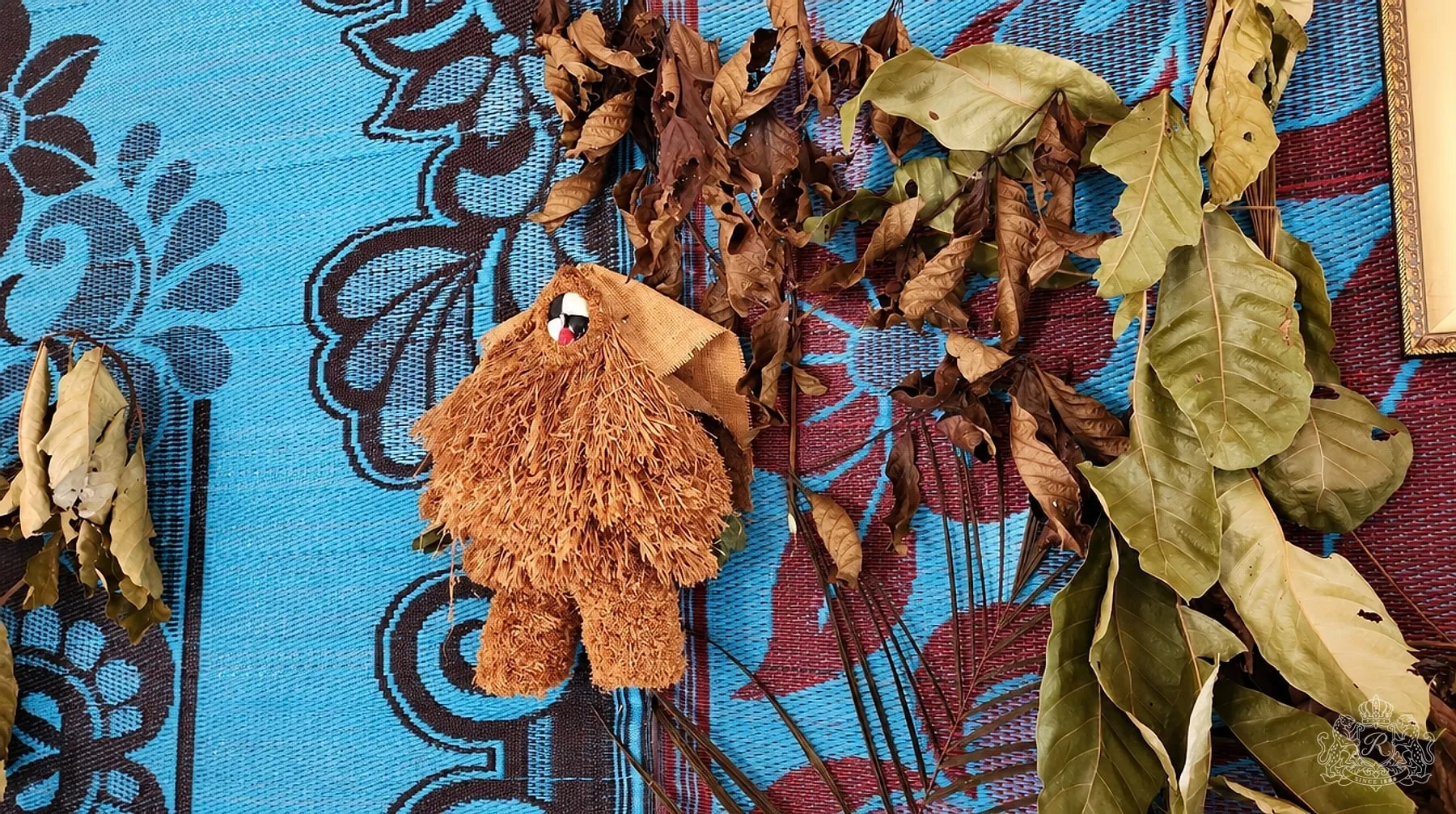 Petite figure de raphia au masque peint posée sur une natte bleue à motifs, entourée de feuilles rituelles séchées
