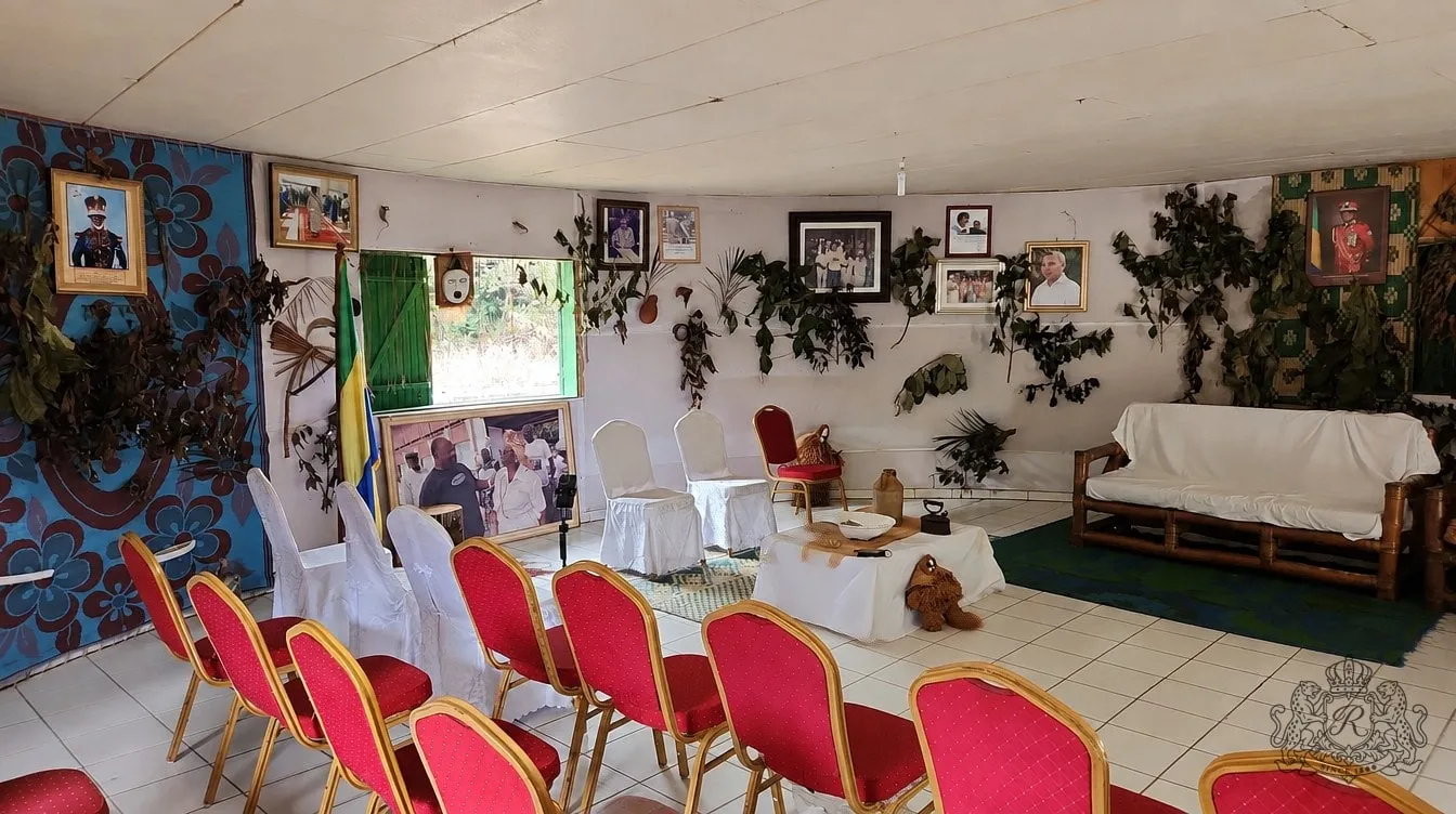 Salle intérieure du musée : portraits encadrés, guirlandes de feuilles séchées, drapeau gabonais, chaises rouges et blanches, table aux objets rituels